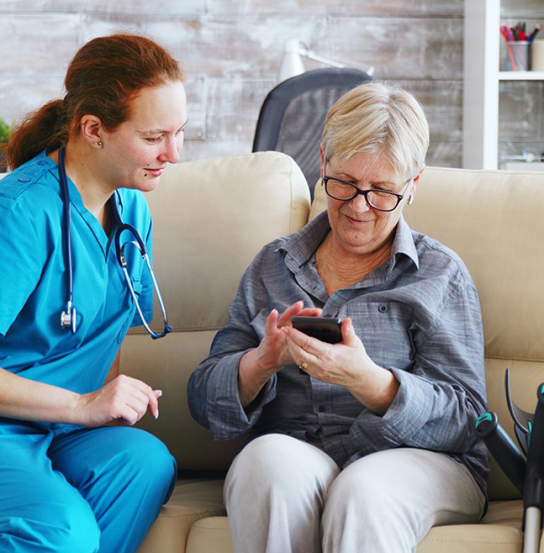 Female doctor in nursing home sitting on the couch teaching senior woman to use smartphone