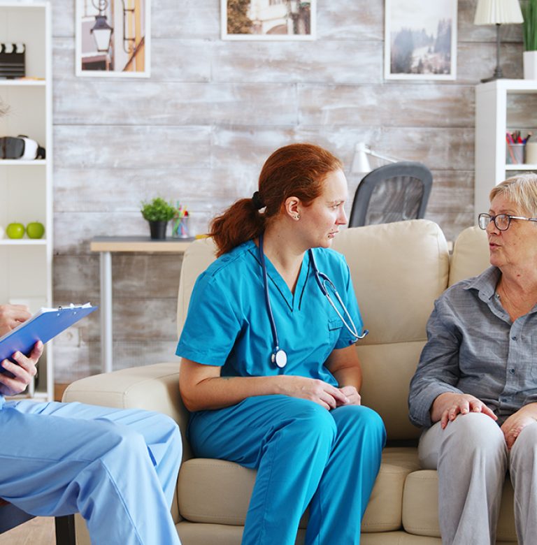 Male assistant taking notes on clipboard while female doctor is talking with senior woman in nursing home